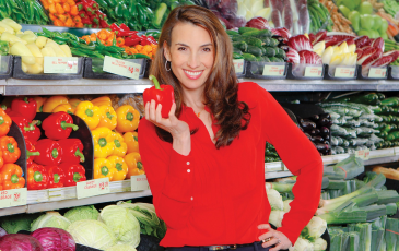 Jessica Siegel in a red blouse holding a red bell pepper behind a grocery store refrigerated vegetable aisle.