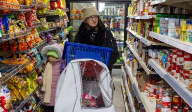 A woman and child walk through the aisles of a grocery store. Several food packages are visible on both sides of them.